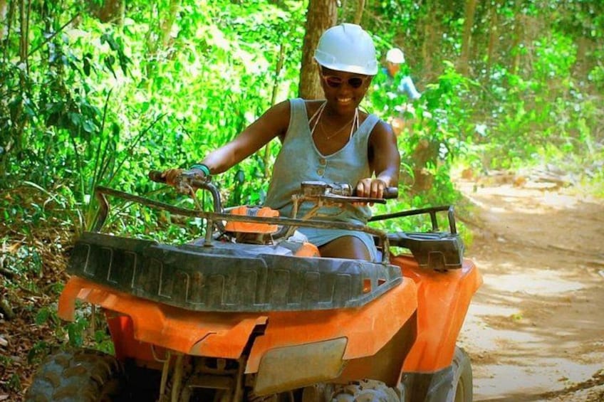 A person riding an orange ATV on a dirt path surrounded by dense green foliage. 1