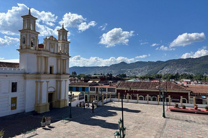 Una plaza con una iglesia, una fuente y montañas al fondo. 1