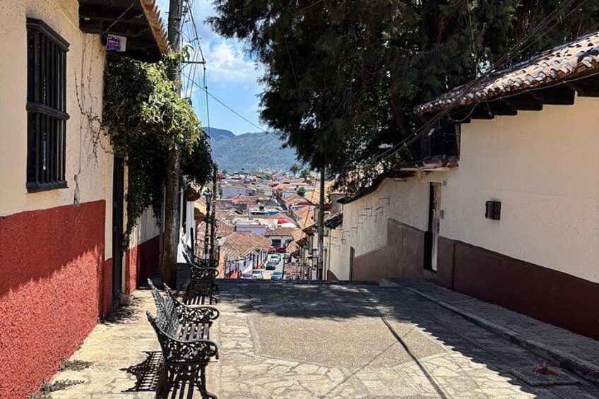 Una calle estrecha flanqueada por edificios, con un banco y una vista distante de un pueblo. 3