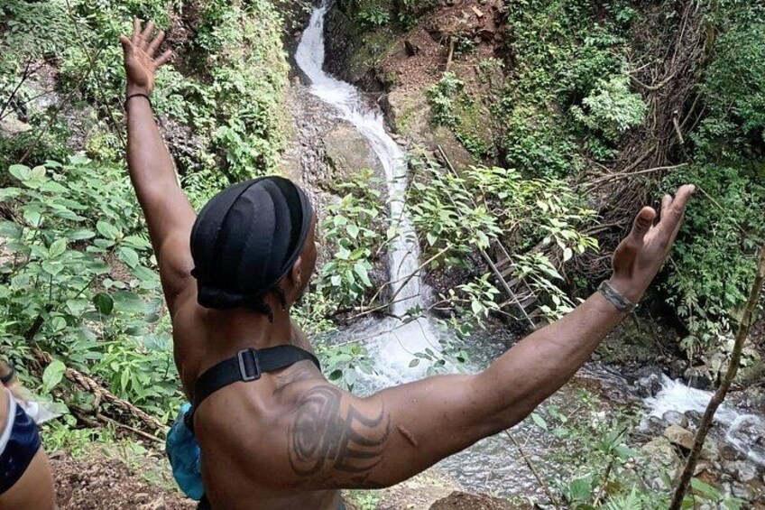 A person standing on a rock with arms raised, overlooking a waterfall in a forested area. 3