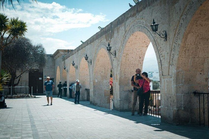 An outdoor walkway with stone arches, a person walking, and another taking a photo. 1