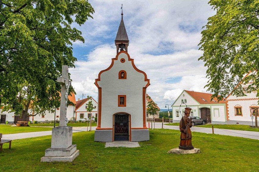 A small church with a bell tower, a statue, and a gravestone in a grassy area with trees and houses in the background. 2