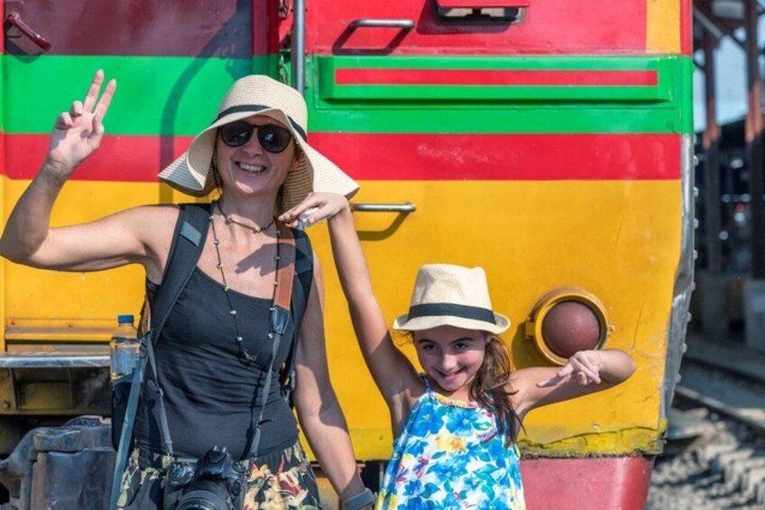 A person in a black tank top and a child in a blue floral dress standing in front of a colorful train. 3