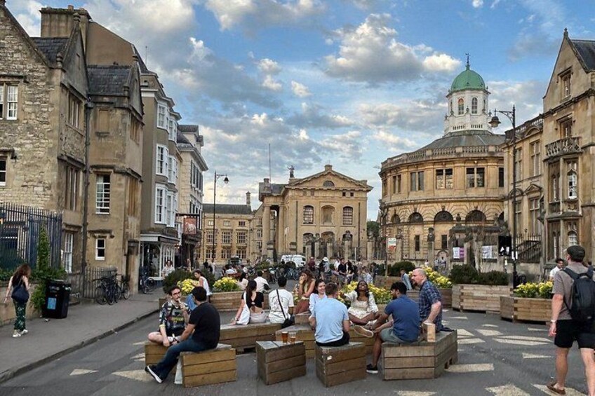 A public square with people sitting on wooden benches, historic buildings, and a prominent church with a green dome. 4