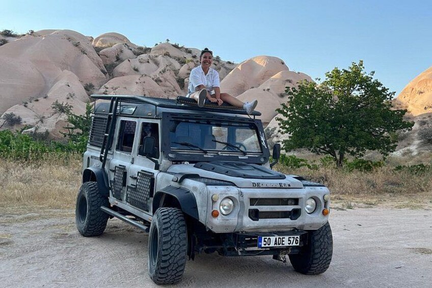 A white off-road vehicle with a person sitting on the roof, parked on a dirt road with large rock formations and sparse vegetation in the background. 4