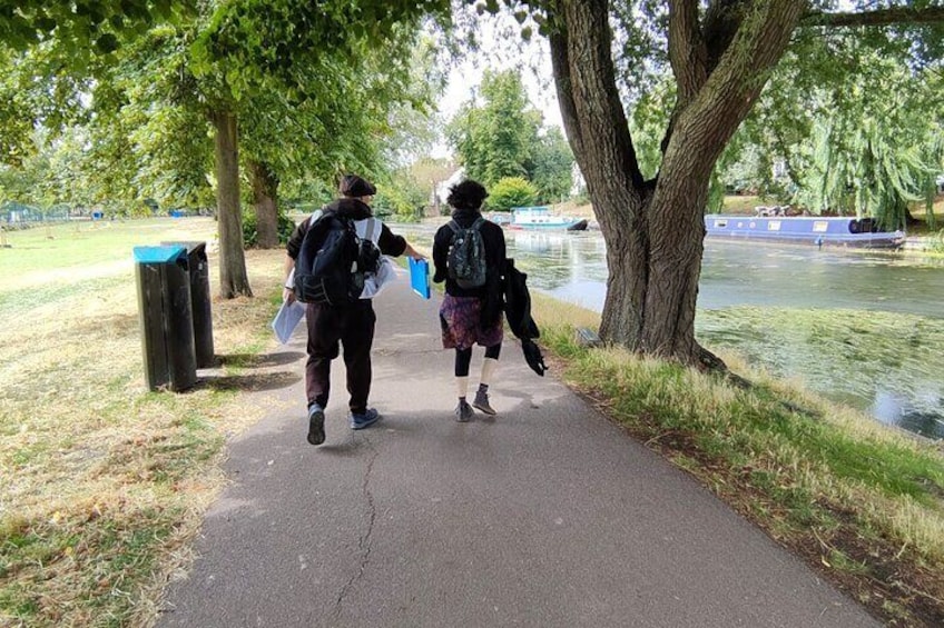 Twee mensen lopen op een pad langs een rivier, met bomen en een prullenbak op de voorgrond. 5