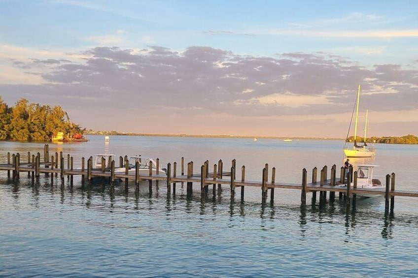 A calm lake with a wooden dock extending into the water, a sailboat anchored near the shore, and a small island with trees in the background. 1