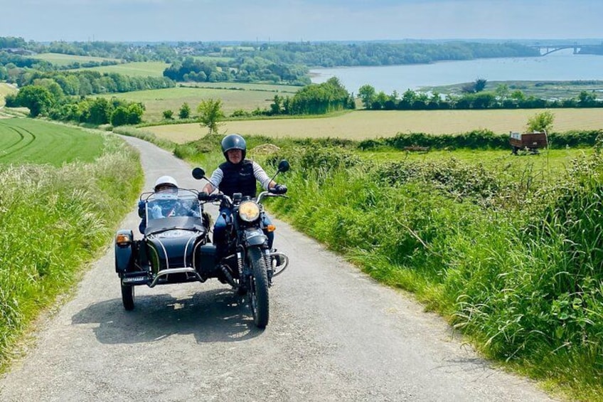 A motorcycle with a sidecar on a rural road with greenery and a bridge in the distance. 2