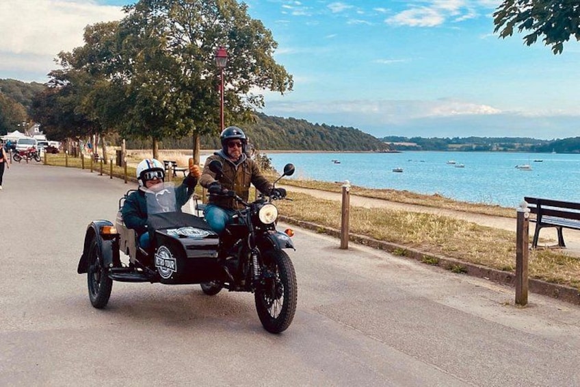 A person riding a motorcycle with a sidecar, another person seated in the sidecar, a lake, boats, and a park with benches. 3