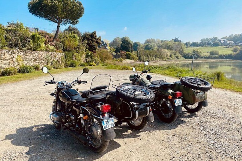 Three ATVs parked on a gravel path by a lake with trees and a stone wall in the background. 3