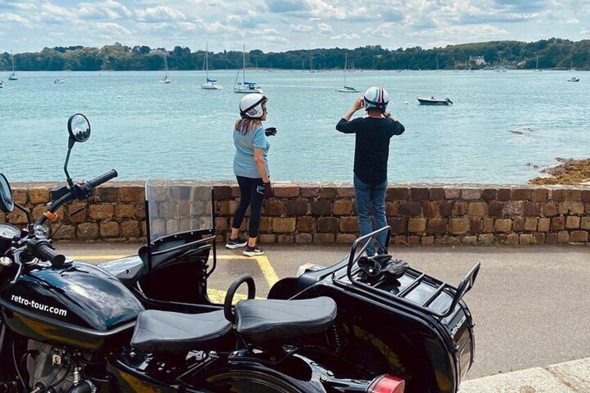 A coastal scene with a stone wall, a parked motorcycle, and two people overlooking the water. 2