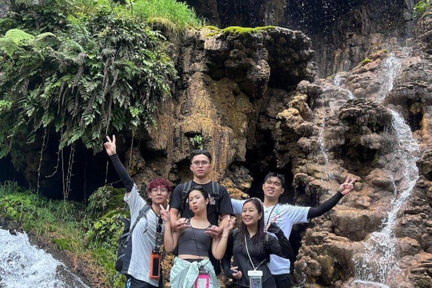 A group of people posing in front of a waterfall with lush greenery and rocky terrain. 3