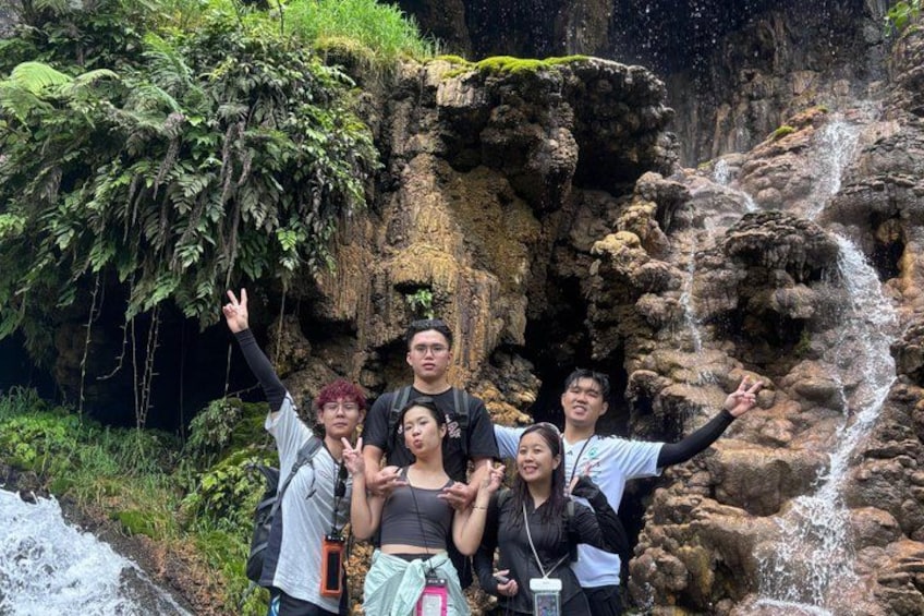 Eine Gruppe von Menschen posiert vor einem Wasserfall, umgeben von üppiger Vegetation und felsigem Gelände. 3