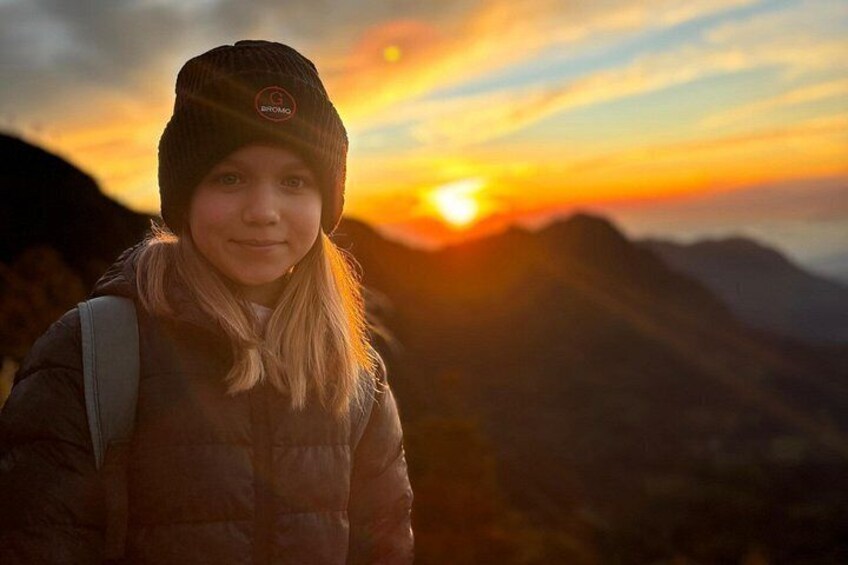 Eine Person mit Mütze und Jacke steht mit einem Rucksack vor einem Sonnenuntergang mit Bergen im Hintergrund. 1