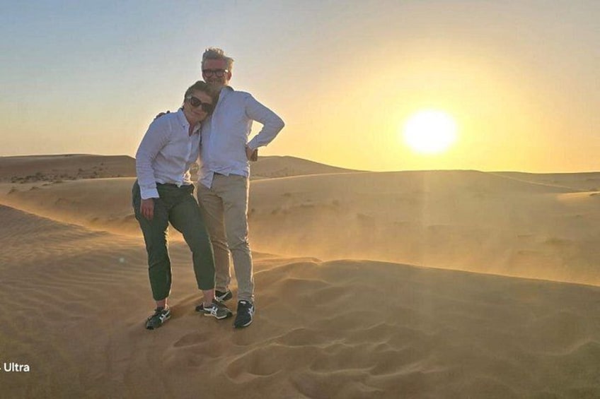 Zwei Personen stehen auf einer Sanddüne vor einer Wüstenlandschaft, während die Sonne untergeht. 1