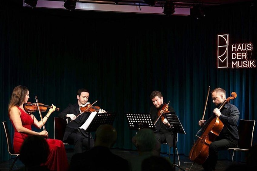 A string quartet performing on stage with music stands and a logo in the background. 1