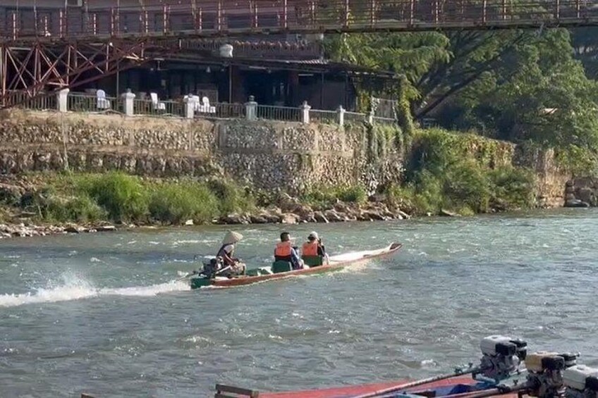 A river with a bridge in the background, a boat with two people, and a rocky shore. 3