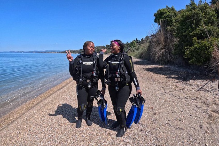 Two divers in black wetsuits with scuba gear on a pebbly beach with clear water and a distant shoreline. 2