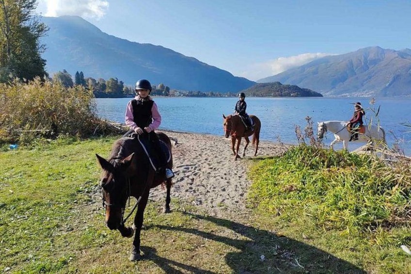 Deux personnes à cheval, qui se promènent le long d’un sentier au bord d’un lac, avec des montagnes en arrière-plan. 1