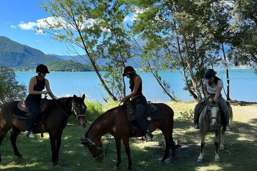Tres personas montando a caballo junto a un lago, con árboles y montañas al fondo. 2