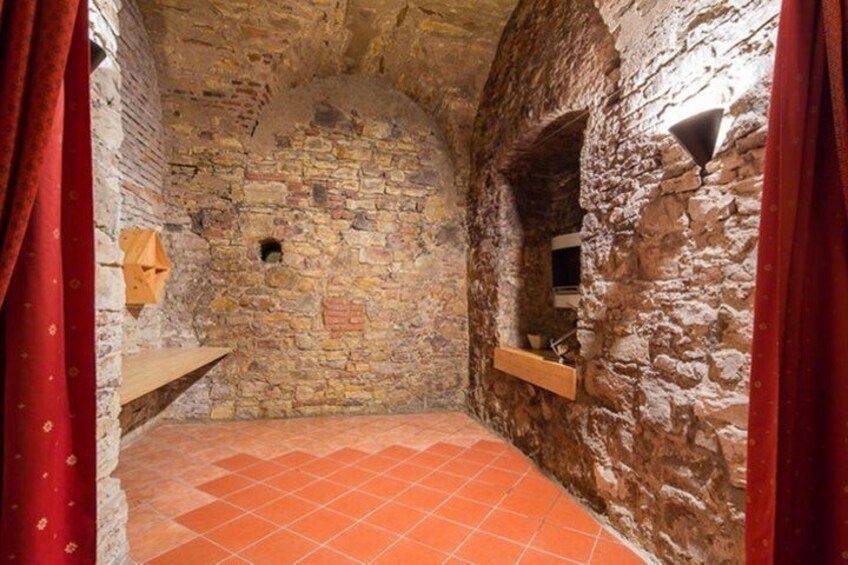 A stone-walled room with a tiled floor, a wooden shelf, and a small window. 3