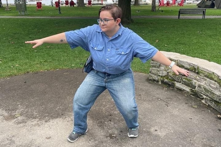A person in a blue shirt and jeans standing on a concrete path in a park with grass and trees in the background. 5