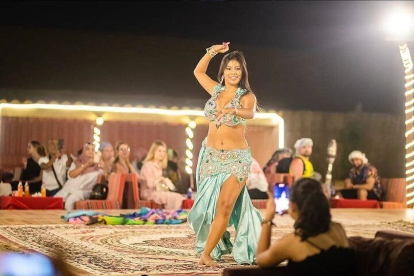 A belly dancer performing on a stage with an audience seated in the background. 3