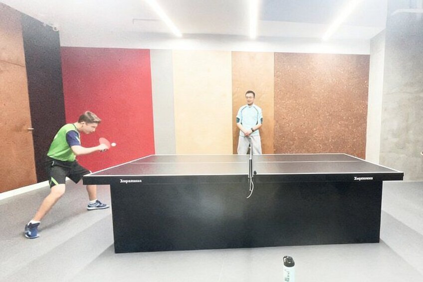 Two people playing table tennis in a room with a ping pong table and walls painted in red and beige. 2