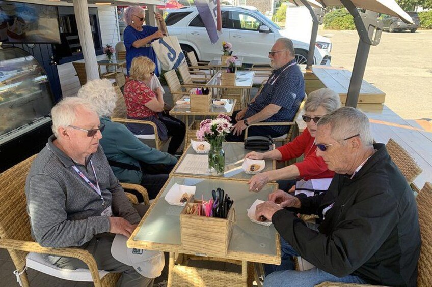 Mensen zitten aan een tafel met eten en drinken, buiten met een auto en een gebouw op de achtergrond. 2