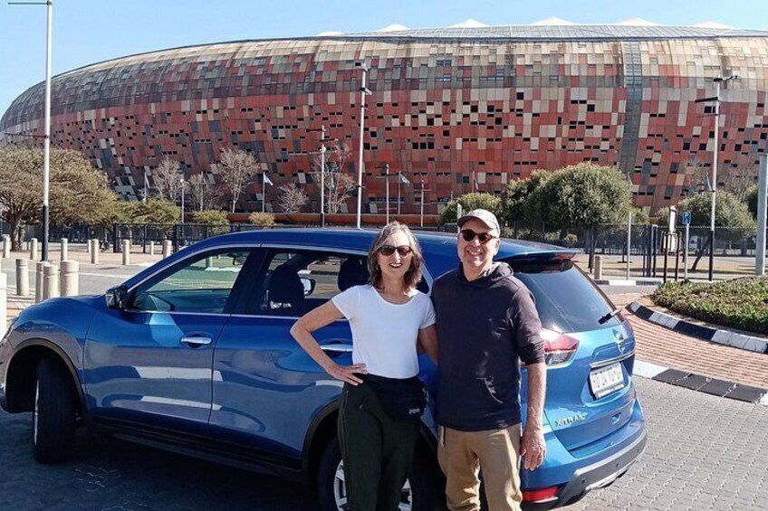 A blue car parked in front of a large, modern stadium with a unique, pixelated facade. 5