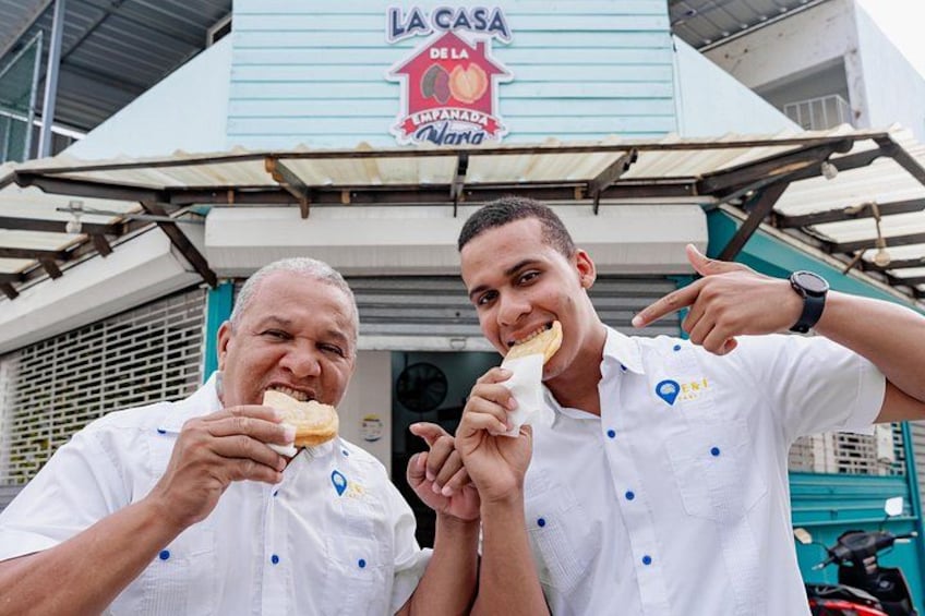 Dos personas con camisas blancas con logotipos, comiendo frente a un edificio con un letrero que dice 'LA CASA DE LA MARA'. 1