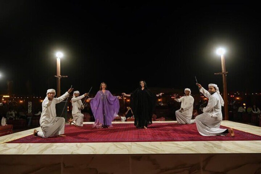 A group performing on a stage with a red carpet, dressed in traditional white attire, under bright stage lights. 1