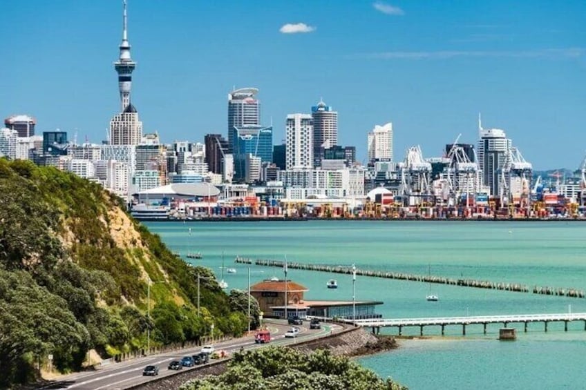 A coastal cityscape with a prominent tower, modern buildings, a bridge, and a clear blue sky. 1