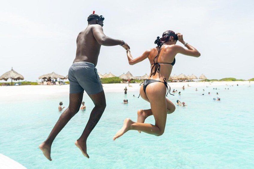 Two people jumping into a clear blue pool at a beach with umbrellas and people in the background. 4