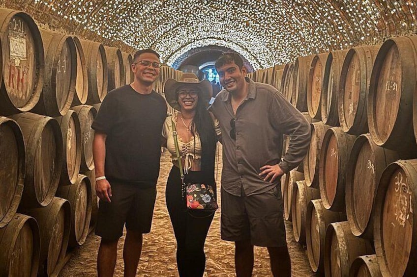 A group of people standing in a wine cellar with rows of wooden barrels. 4