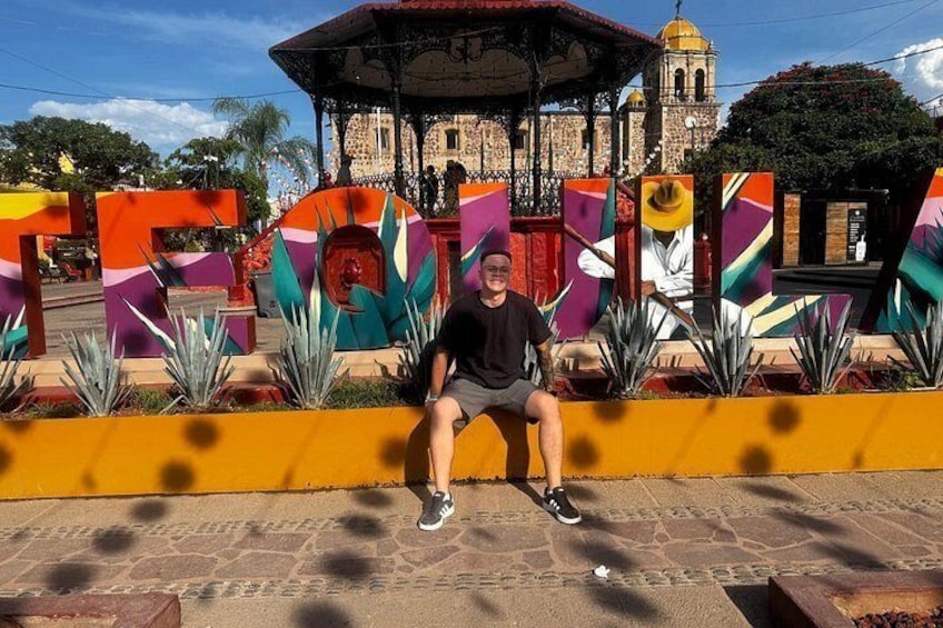 A man sitting on a bench in front of a colorful mural with geometric shapes and a gazebo in the background. 1