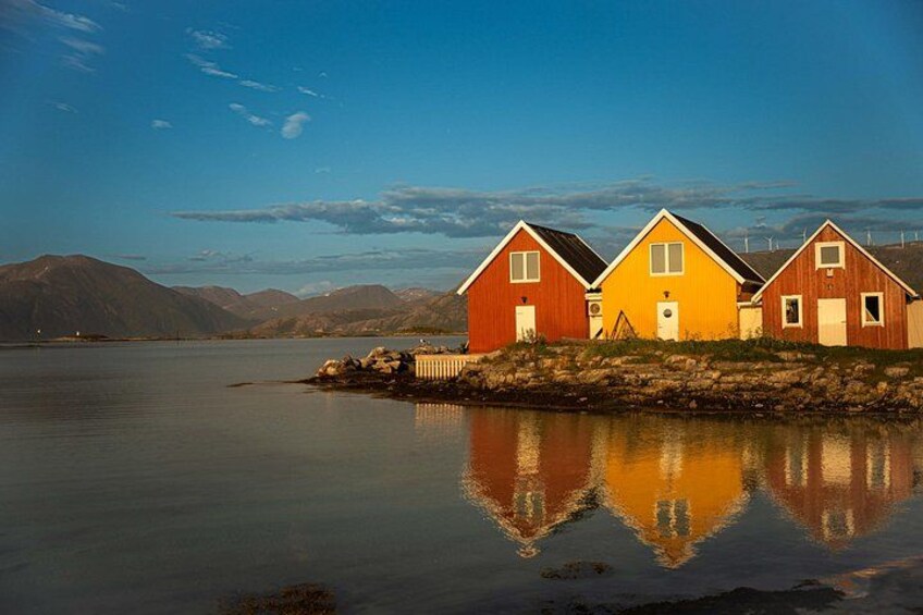 Three wooden houses by a calm lake with mountains in the background. 1