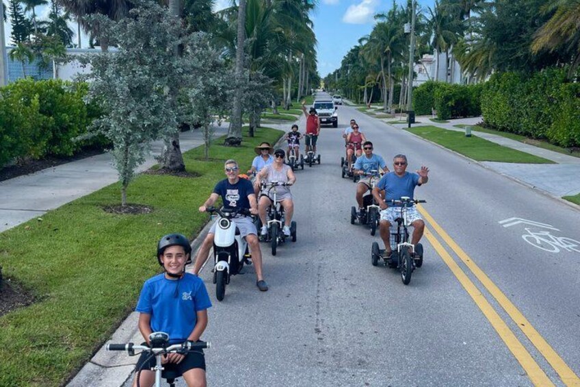 A group of people riding electric scooters on a street lined with palm trees and greenery. 2