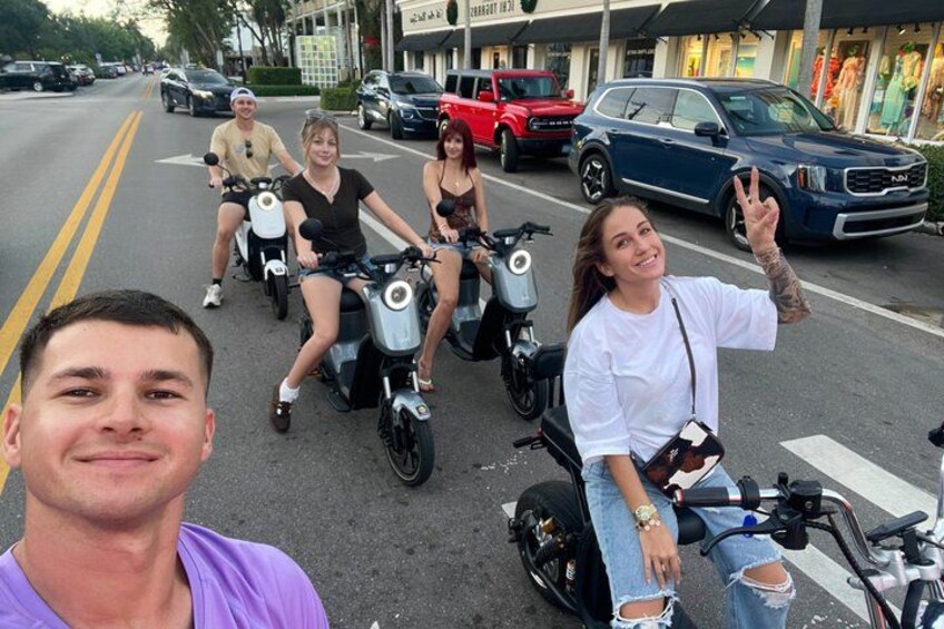 Four people on scooters on a street with cars and buildings in the background. 1