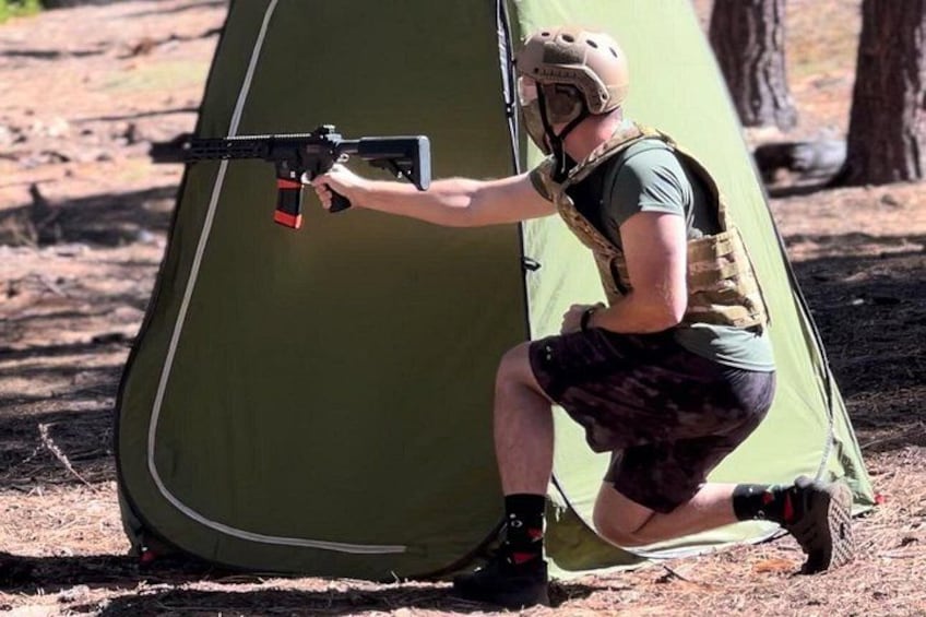 Una persona agachada al lado de una tienda de campaña verde, sosteniendo un rifle. 4