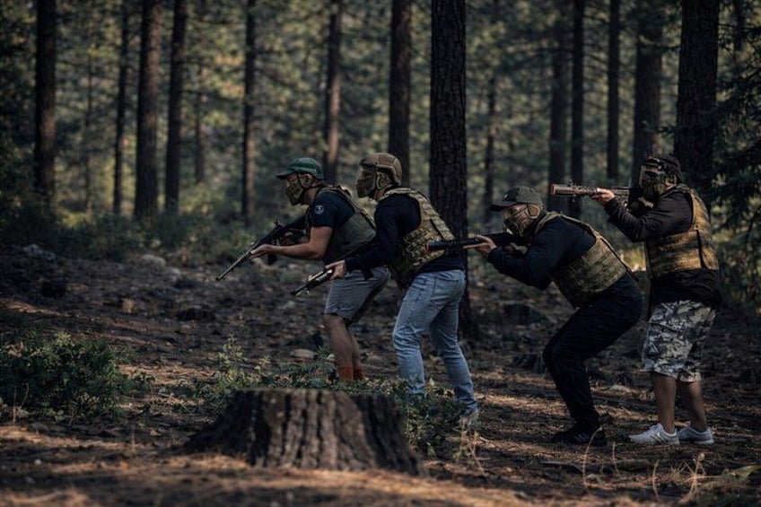 Vier Personen in einem Wald mit Gewehren, ausgestattet mit taktischer Ausrüstung. 1