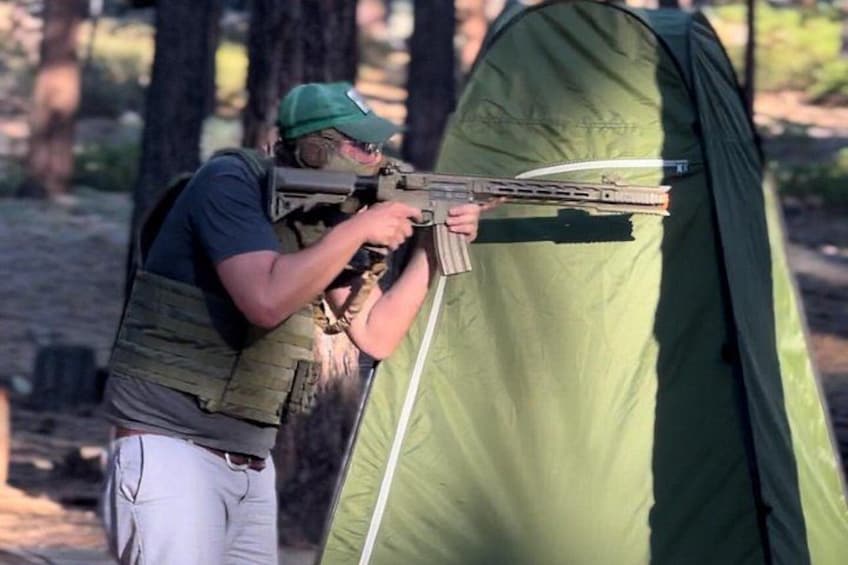Una persona apunta con un rifle a una tienda de campaña verde en un área boscosa. 5