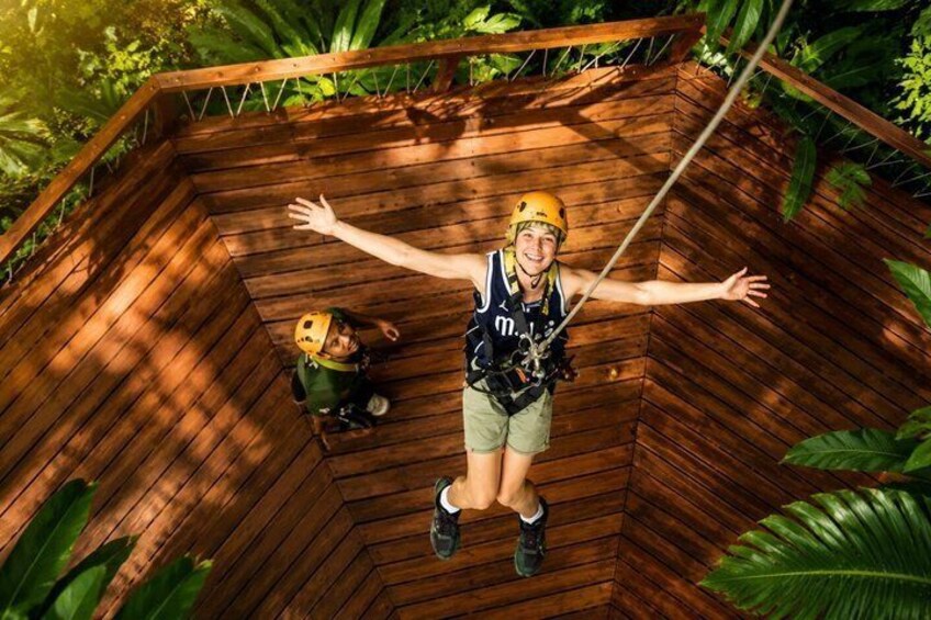 A person on a wooden zipline with a helmet and harness, another person sitting on the ground, surrounded by greenery. 4
