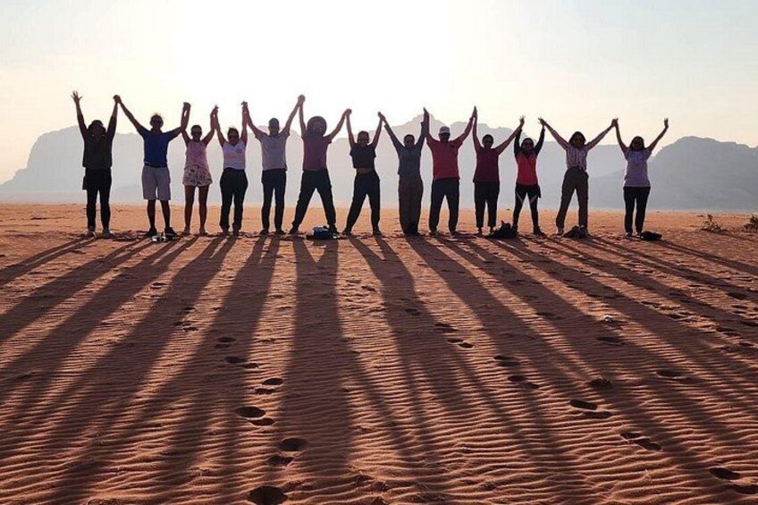 A group of people standing in a line on a sandy beach with their arms raised, facing a mountainous landscape. 2
