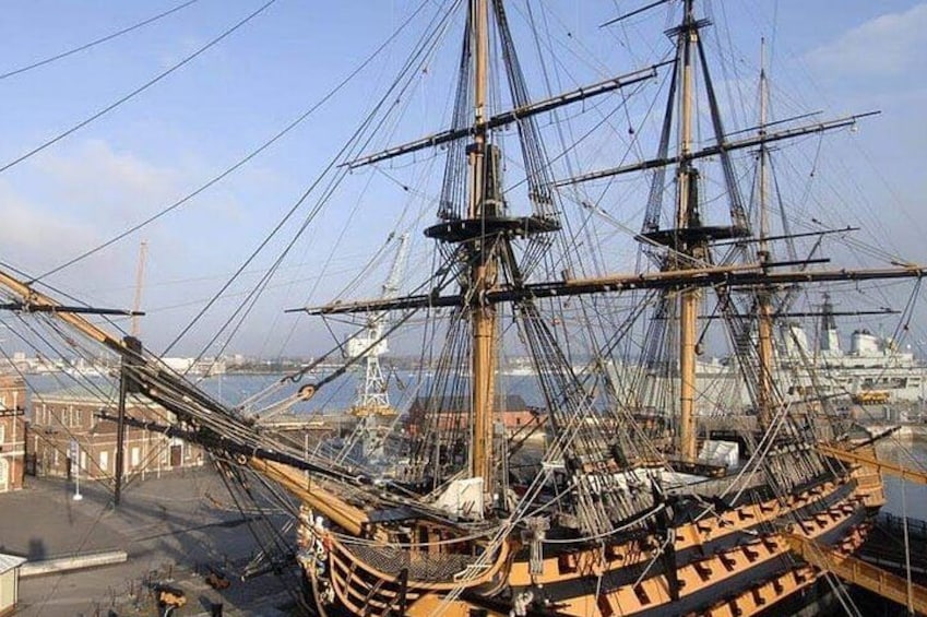 Ein großes Segelschiff liegt im Hafen festgemacht, mit mehreren Masten und Rigg, umgeben von anderen Schiffen und Gebäuden. 4