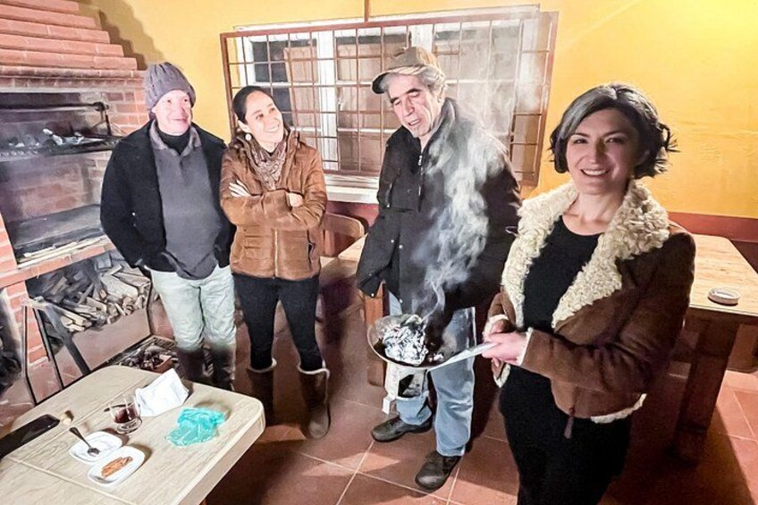 Persone intorno a un forno a legna, con fumo che si alza, e un tavolo imbandito di cibo e bevande. 2