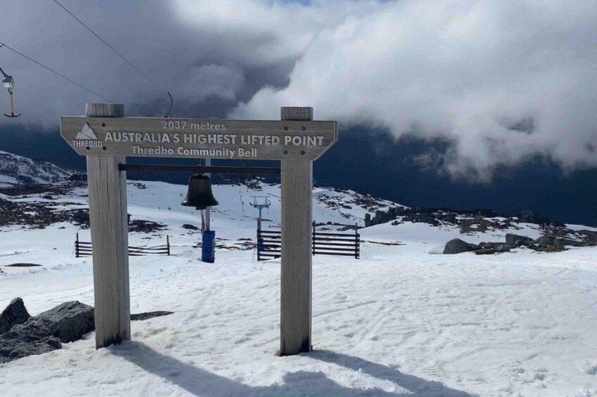 A sign indicating Australia's highest lifted point at 2037 meters, Thredbo Community Bell, with a snowy landscape and a ski lift in the background. 3