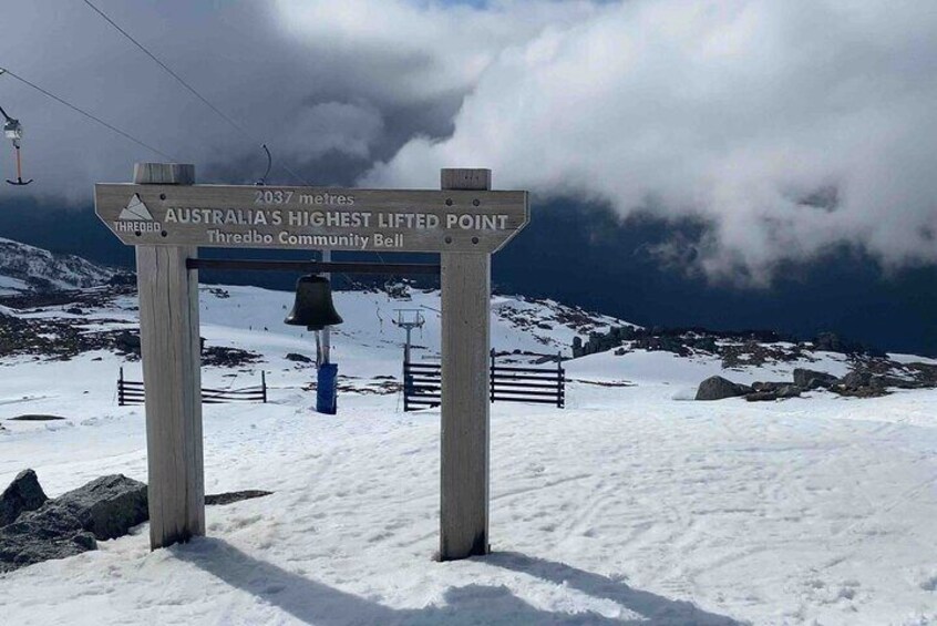 A sign marking Australia's highest lifted point at 2037 meters, Thredbo Community Bell, with a snowy landscape and a ski lift in the background. 3