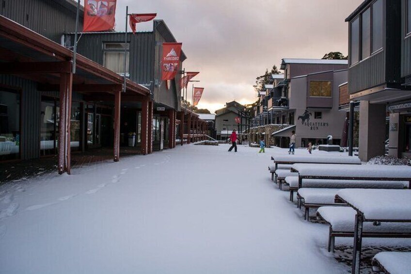 A snow-covered street with buildings and benches. 5