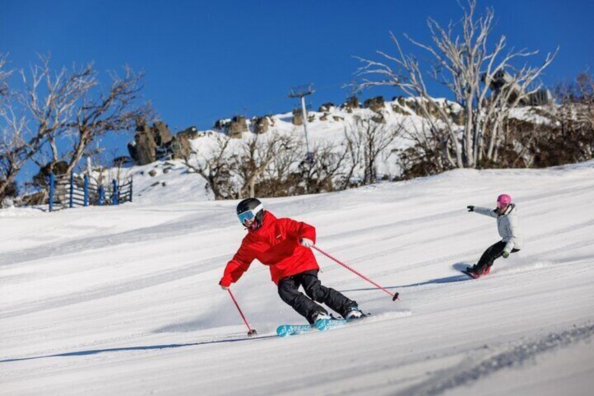 Two people skiing down a snowy slope with a clear blue sky and rocky terrain in the background. 4